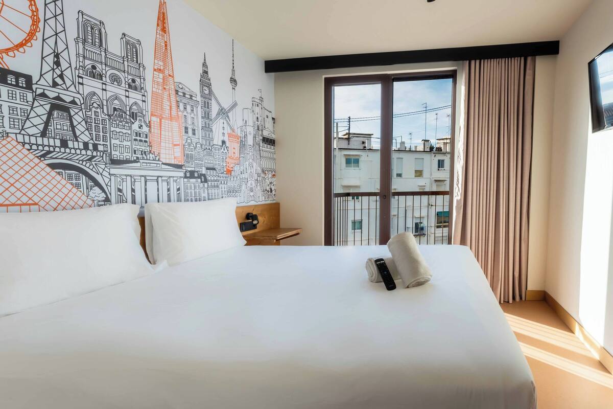 A hotel room with a large bed, a nightstand, a wall mural of a cityscape, and a large window with curtains.