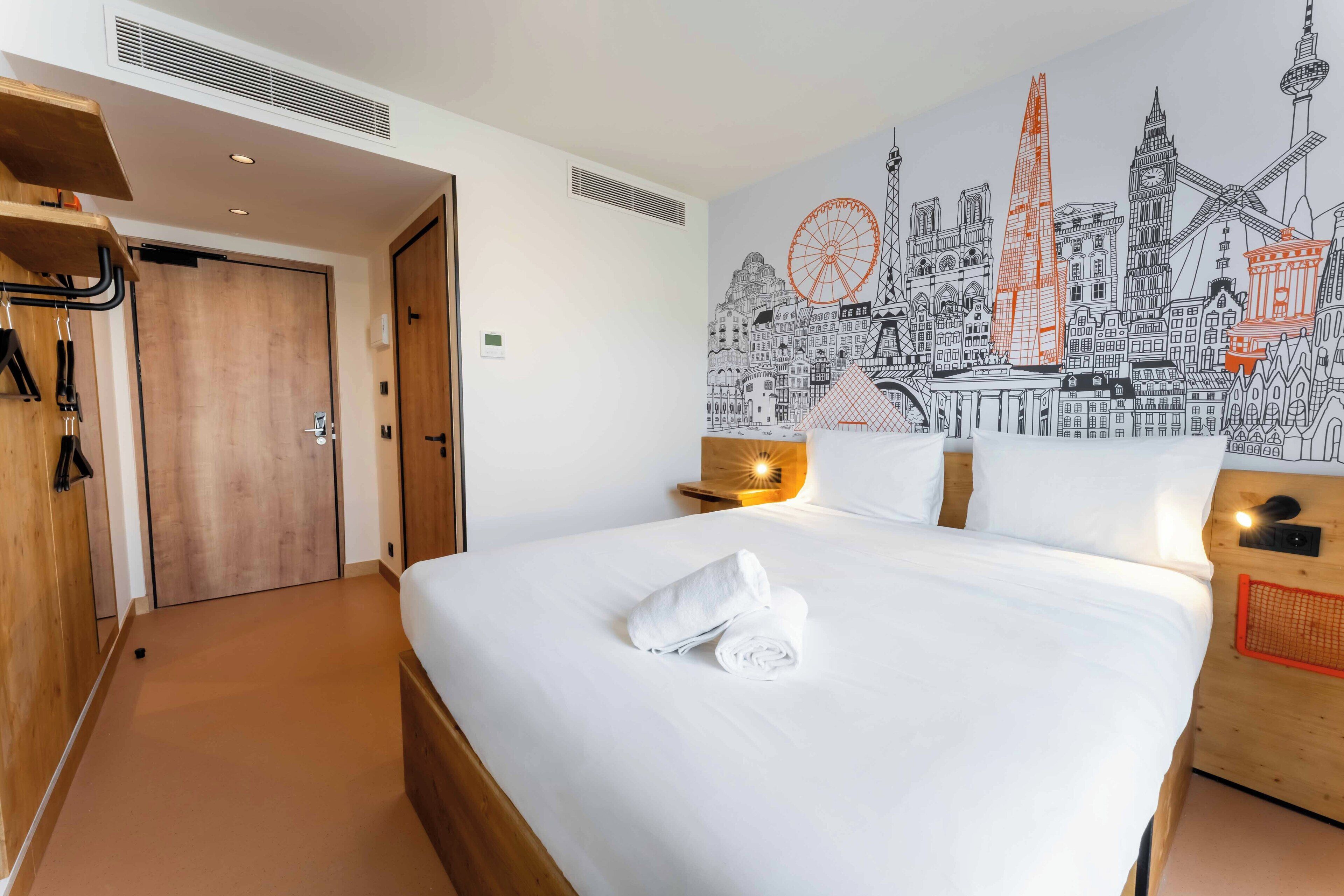 Una habitación de hotel con una cama grande, un armario de madera, un mural en la pared con vista a un paisaje urbano y una mesita con una lámpara.