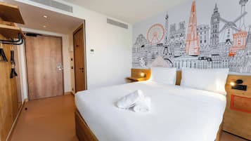 Una habitación de hotel con una cama grande, un armario de madera, un mural en la pared con vista a un paisaje urbano y una mesita con una lámpara.