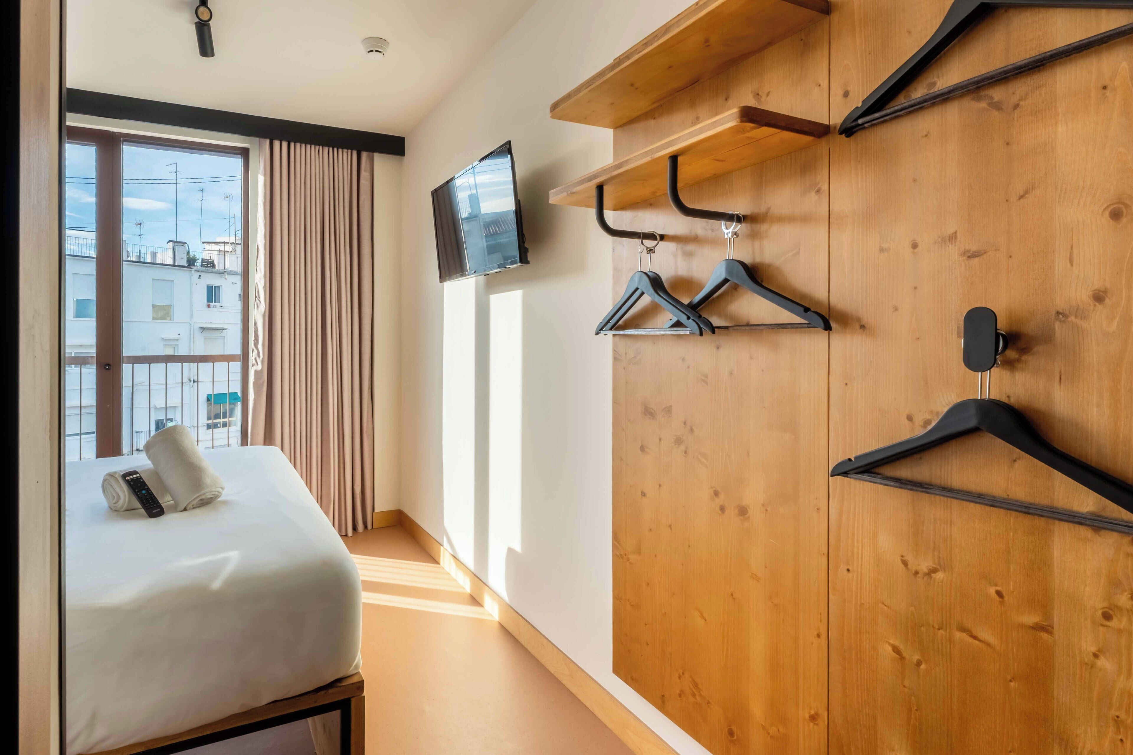 Habitación de hotel con cama, televisor montado en la pared, armario de madera y ventana con cortinas.