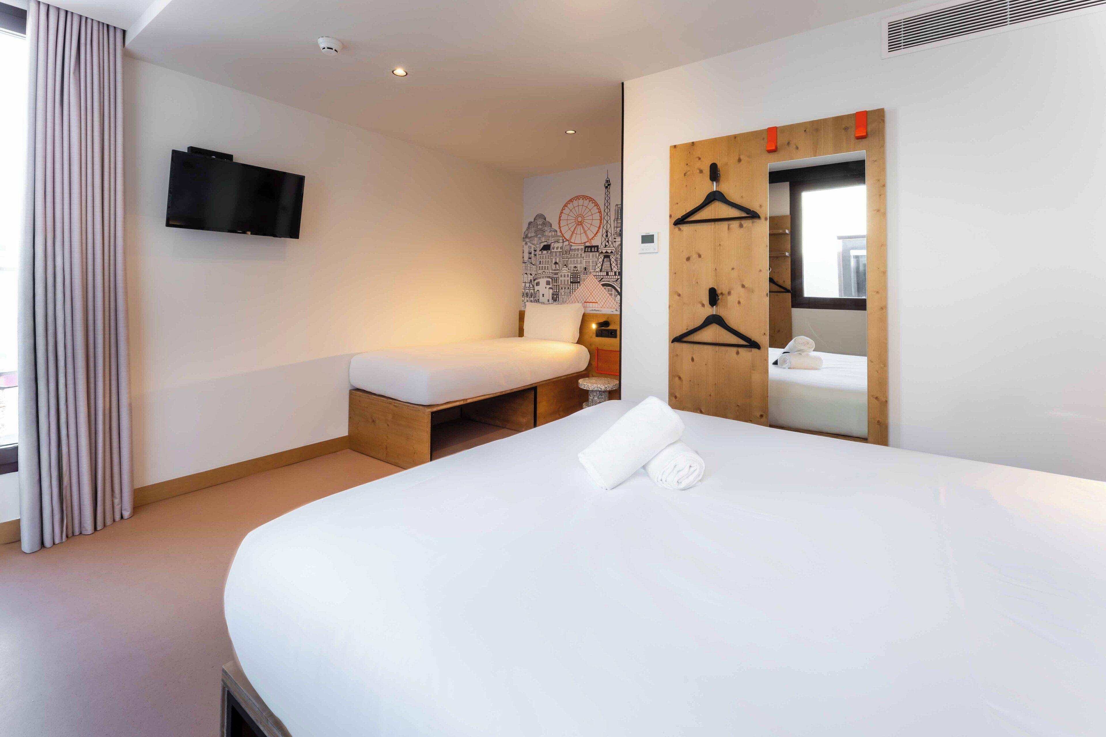 A modern hotel room with a large bed, a smaller bed, a TV mounted on the wall, a wooden wardrobe with a mirror, and a cityscape mural on the wall.