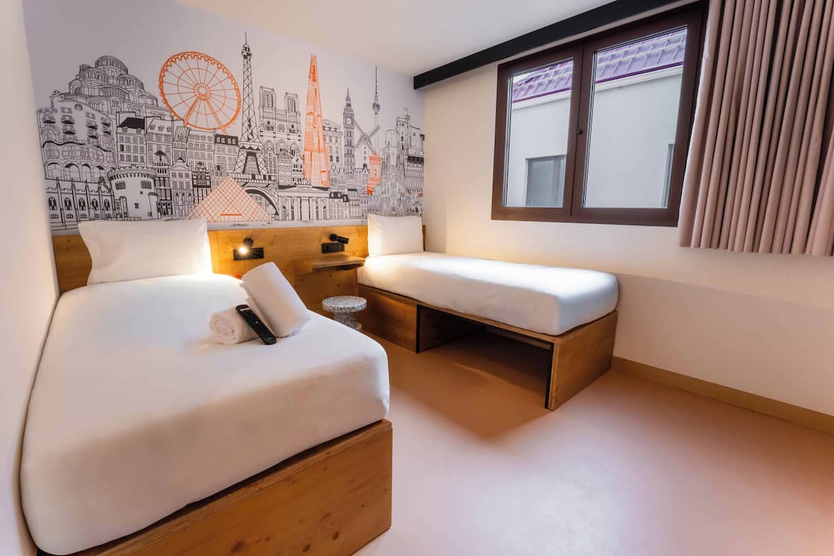 A hotel room with a bed, a small desk, a nightstand, a window with curtains, and a cityscape mural on the wall.
