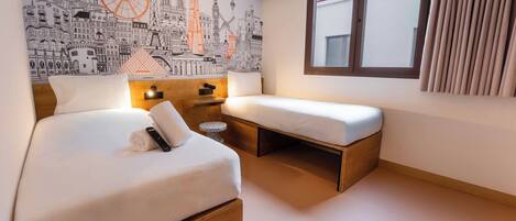 A hotel room with a bed, a small desk, a nightstand, a window with curtains, and a cityscape mural on the wall.