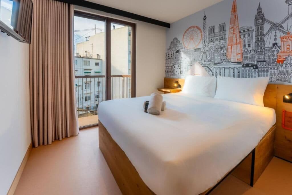 A hotel room with a large bed, a cityscape mural, a window with curtains, and a nightstand with a lamp.