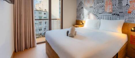 A hotel room with a large bed, a cityscape mural, a window with curtains, and a nightstand with a lamp.