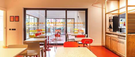 A modern dining area with tables and chairs, a kitchen area with appliances, and large windows allowing natural light.