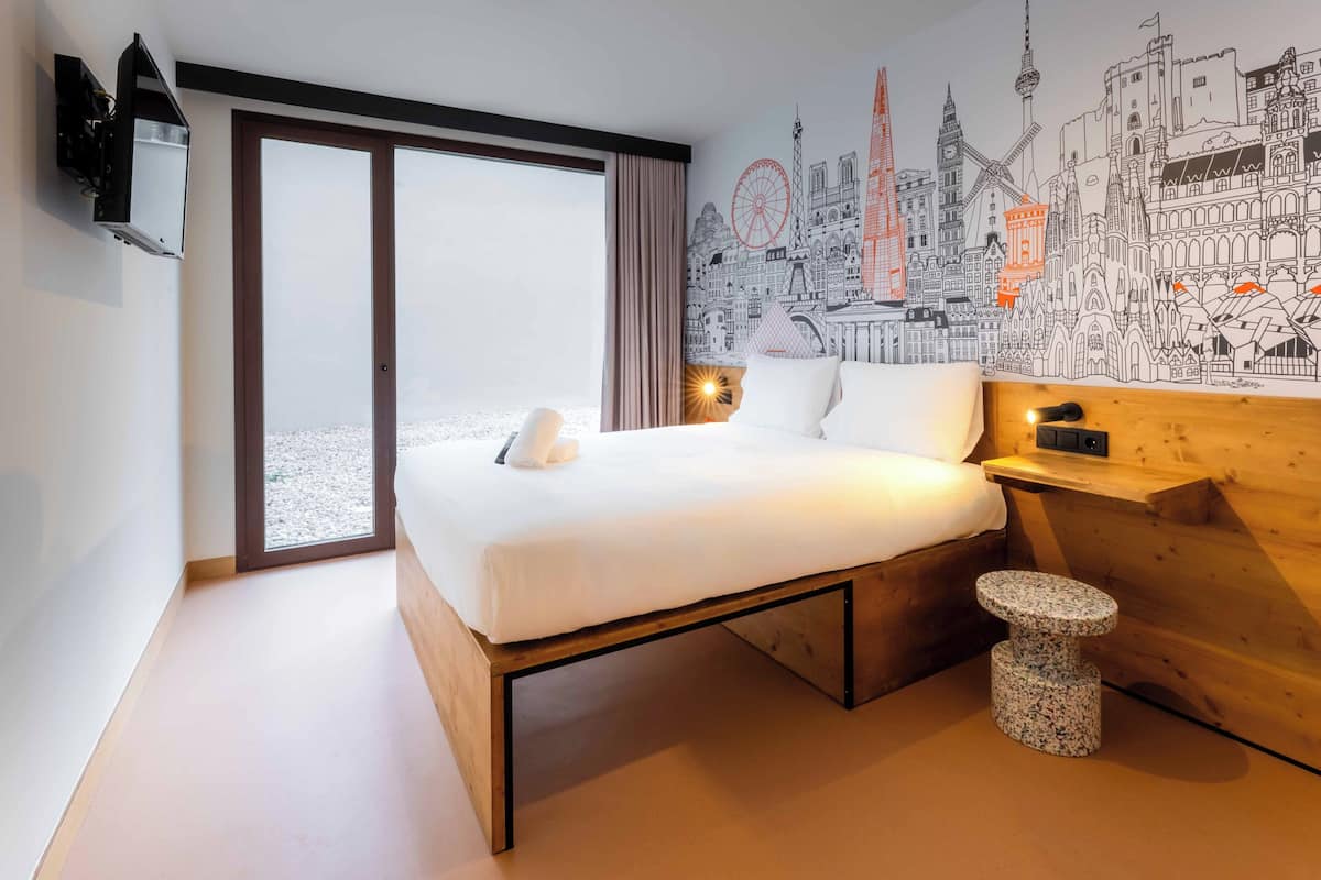 A modern hotel room with a large bed, a wooden nightstand, a small round table, and a cityscape mural on the wall.