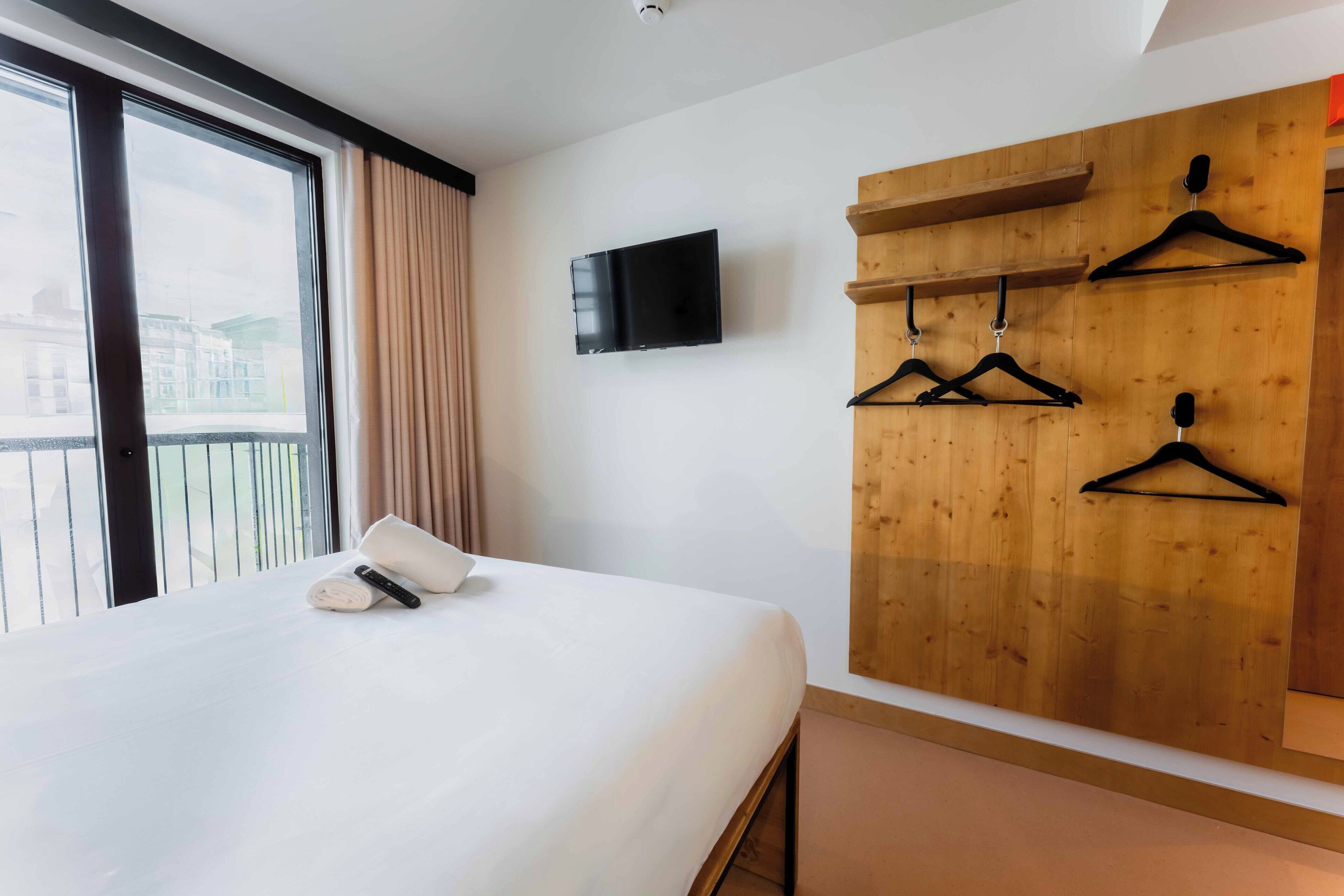 Une chambre d’hôtel avec un grand lit, un téléviseur fixé au mur et des panneaux muraux en bois équipés de porte-manteaux.
