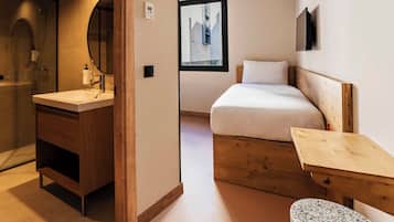 A hotel room with a wooden bed, a small wooden table, a bench, a bathroom with a mirror and sink, and a window with a view of a building.