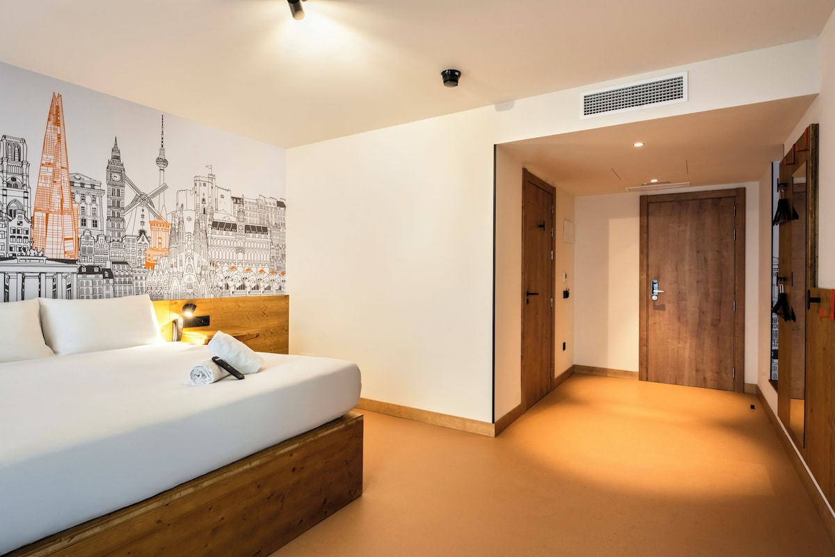 A hotel room with a large bed, a cityscape mural, a wooden headboard, and a hallway leading to a door.