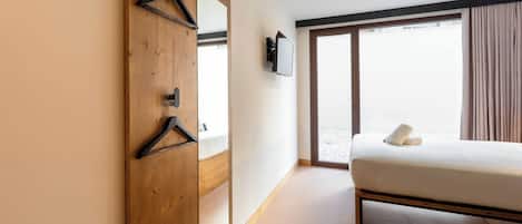 A bedroom with a bed, a wooden wardrobe, a mirror, a TV, and a large window with curtains.