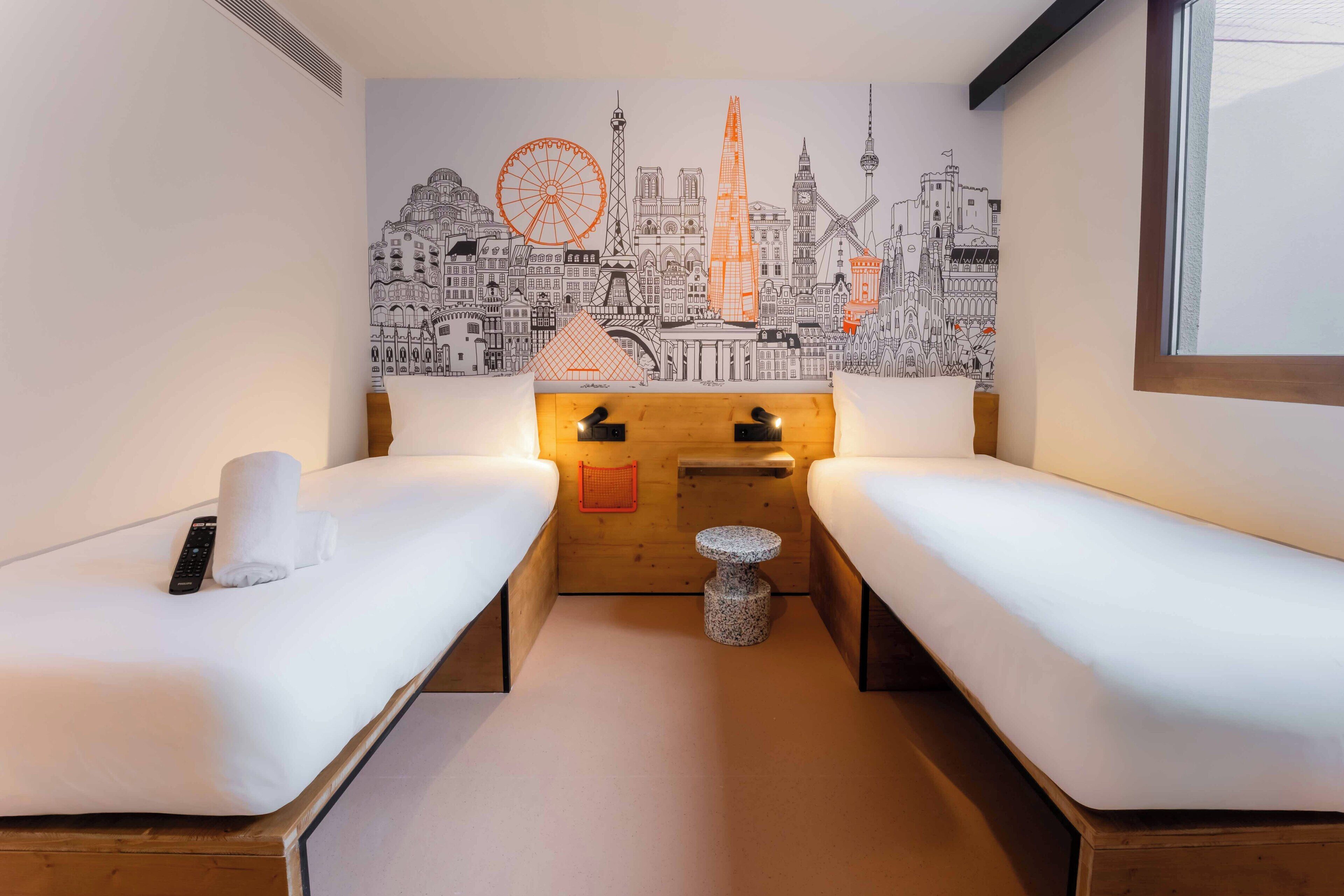A hotel room with two beds, a wooden nightstand, a small round table, and a cityscape mural on the wall.