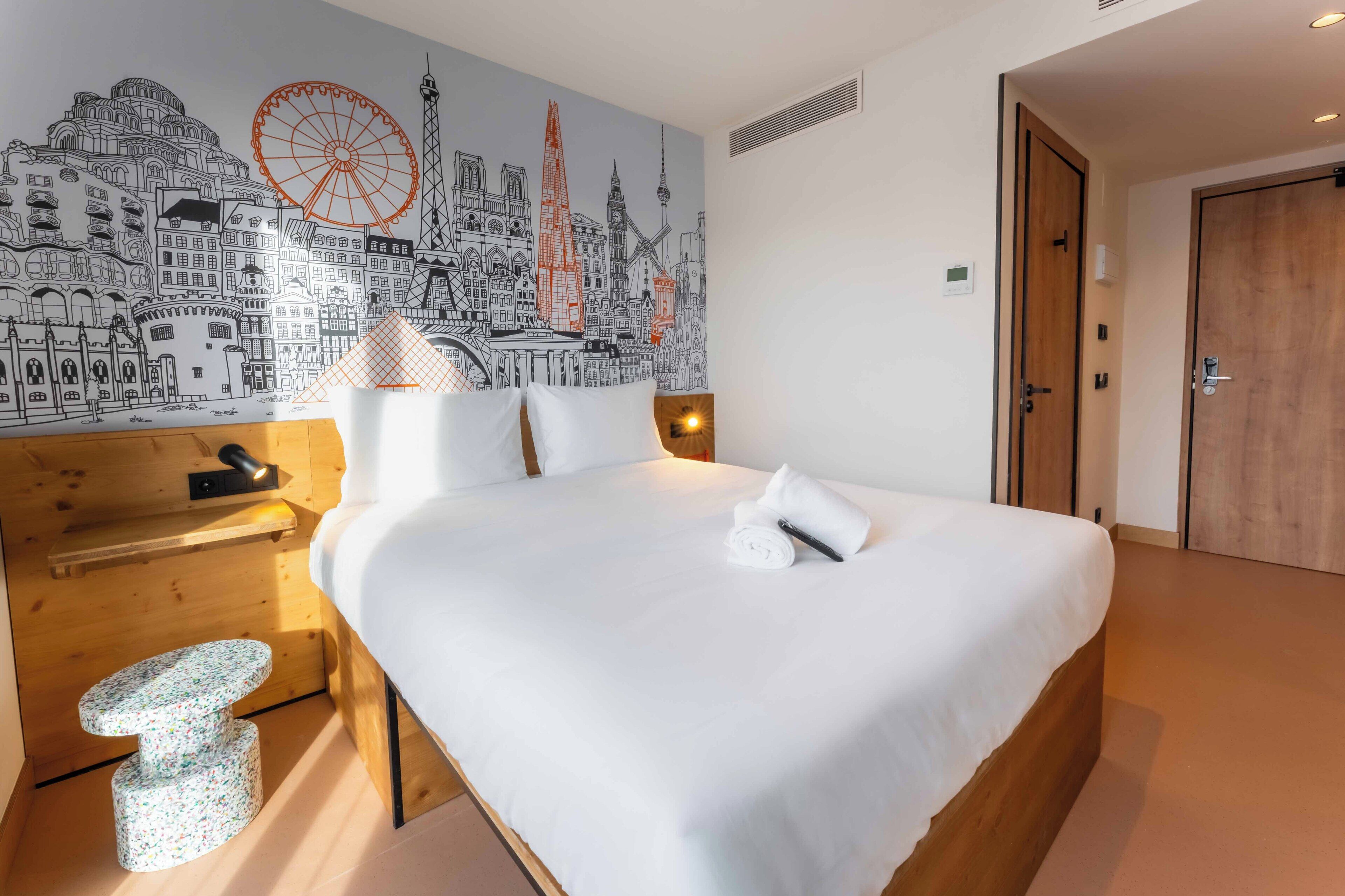 Una camera d'hotel con un letto, un comodino in legno, una lampada, un murale raffigurante un panorama cittadino e una porta.