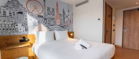 Una habitación de hotel con cama, mesita de noche de madera, lámpara, mural en la pared con vista urbana y una puerta.