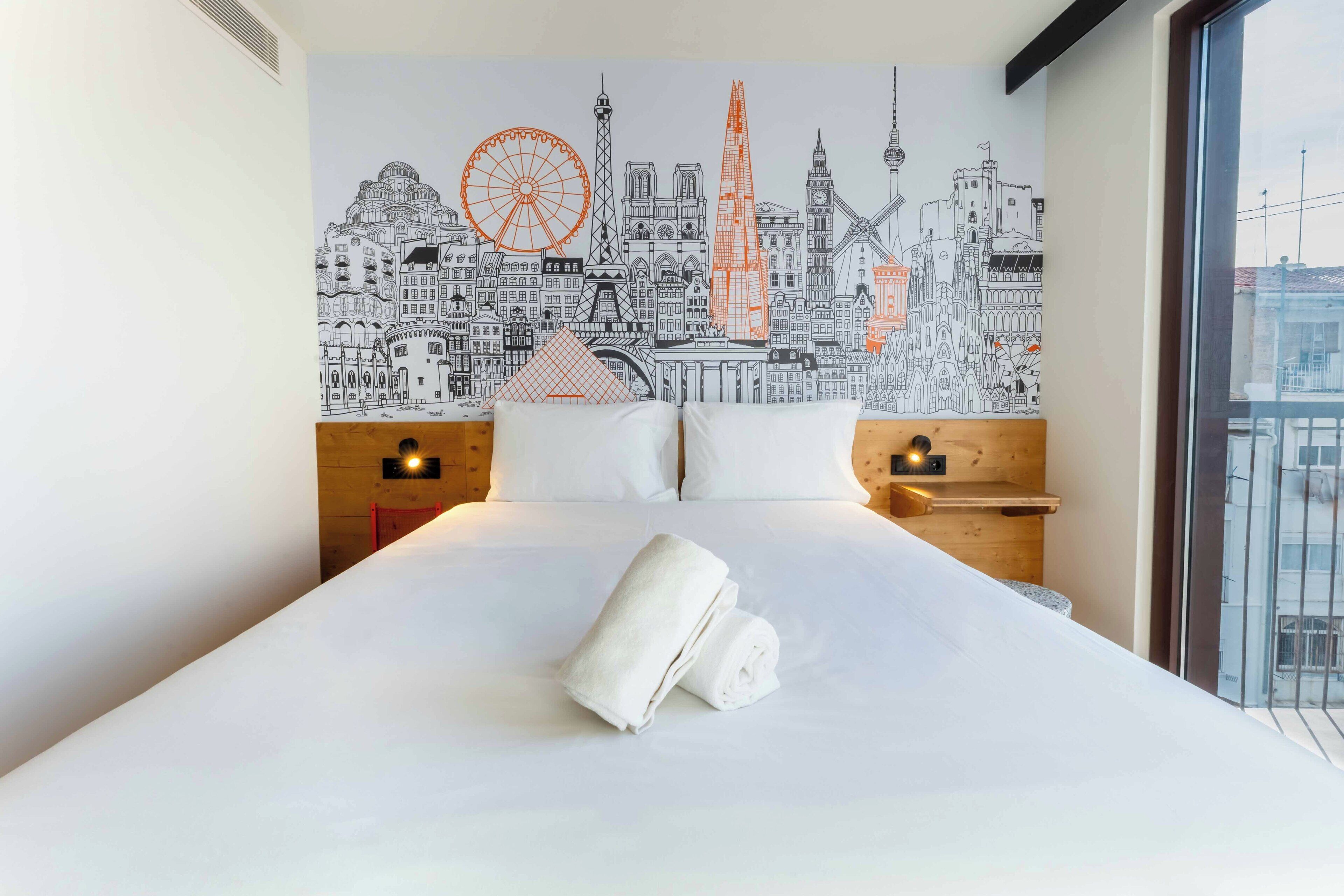 Habitació d'hotel amb un llit, roba de llit blanca, un mural de paisatge urbà a la paret i una finestra amb vistes.
