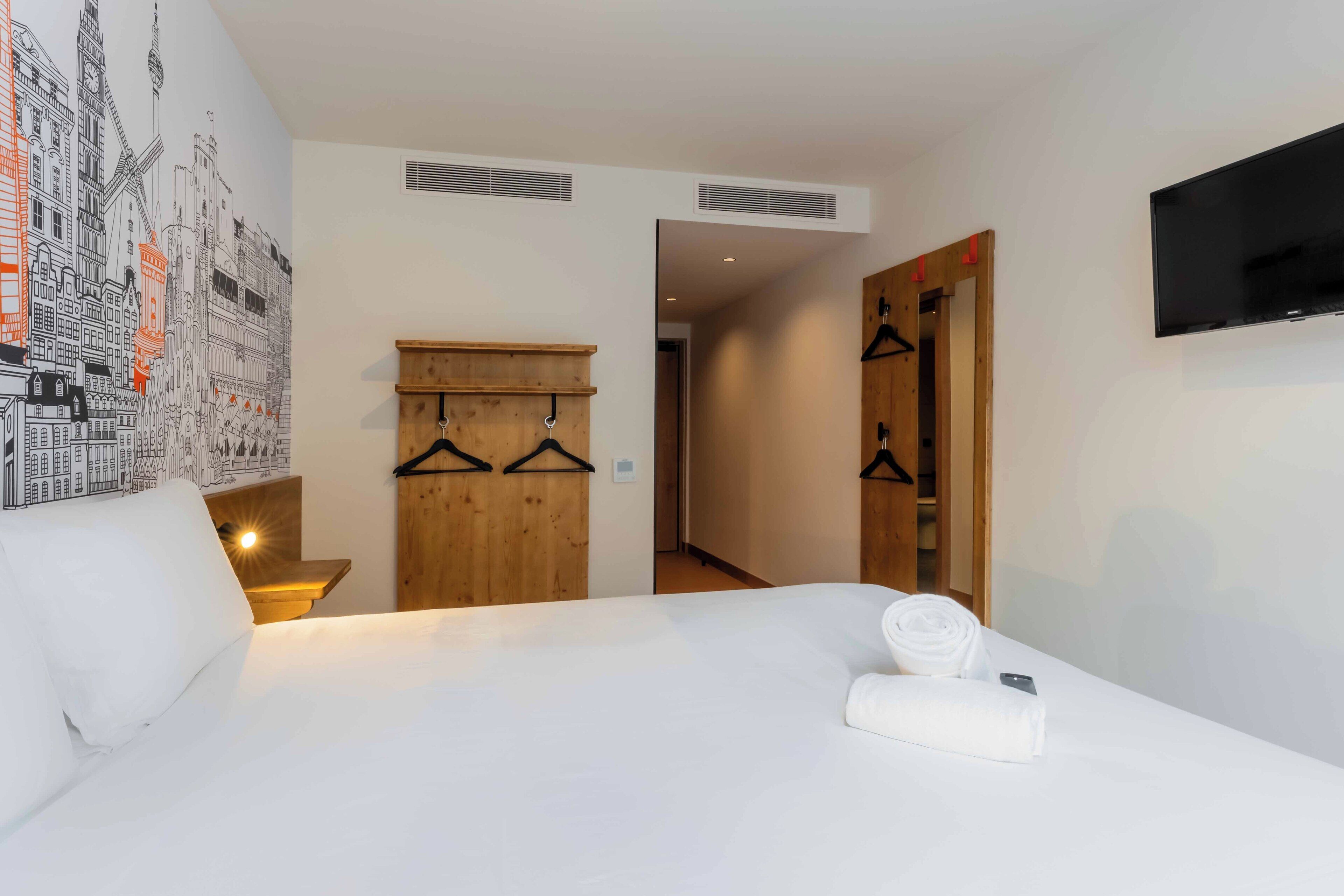 A hotel room with a bed, a wooden wardrobe, a wall-mounted TV, and a cityscape mural.