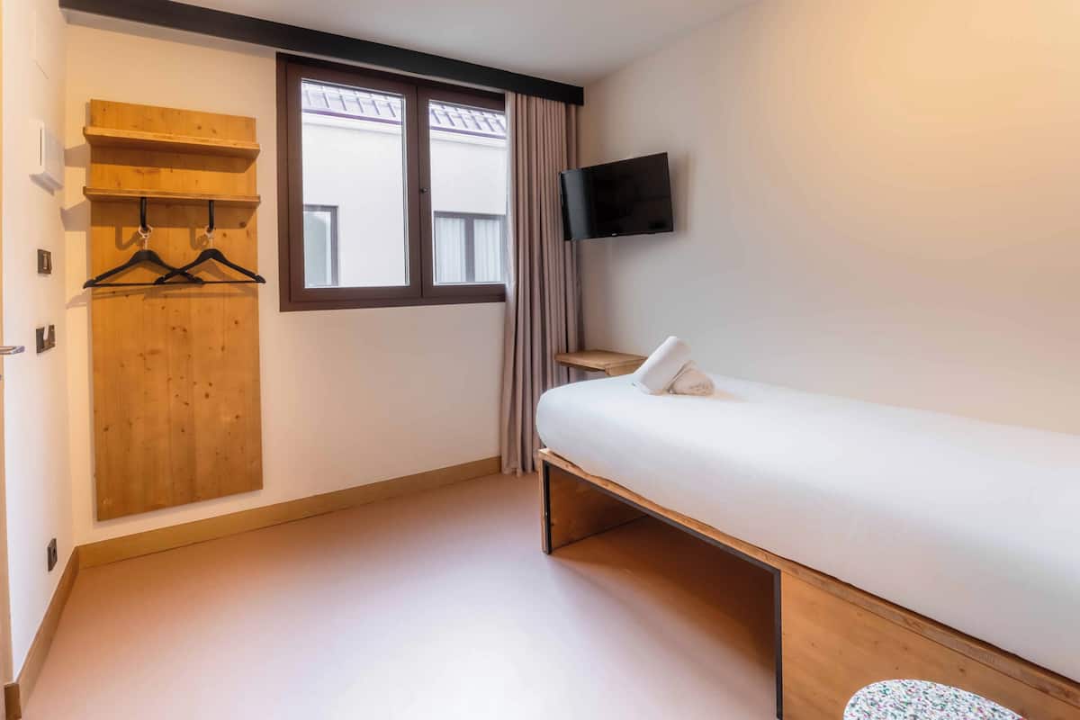 A bedroom with a bed, a TV mounted on the wall, a wooden wardrobe, and a window with curtains.