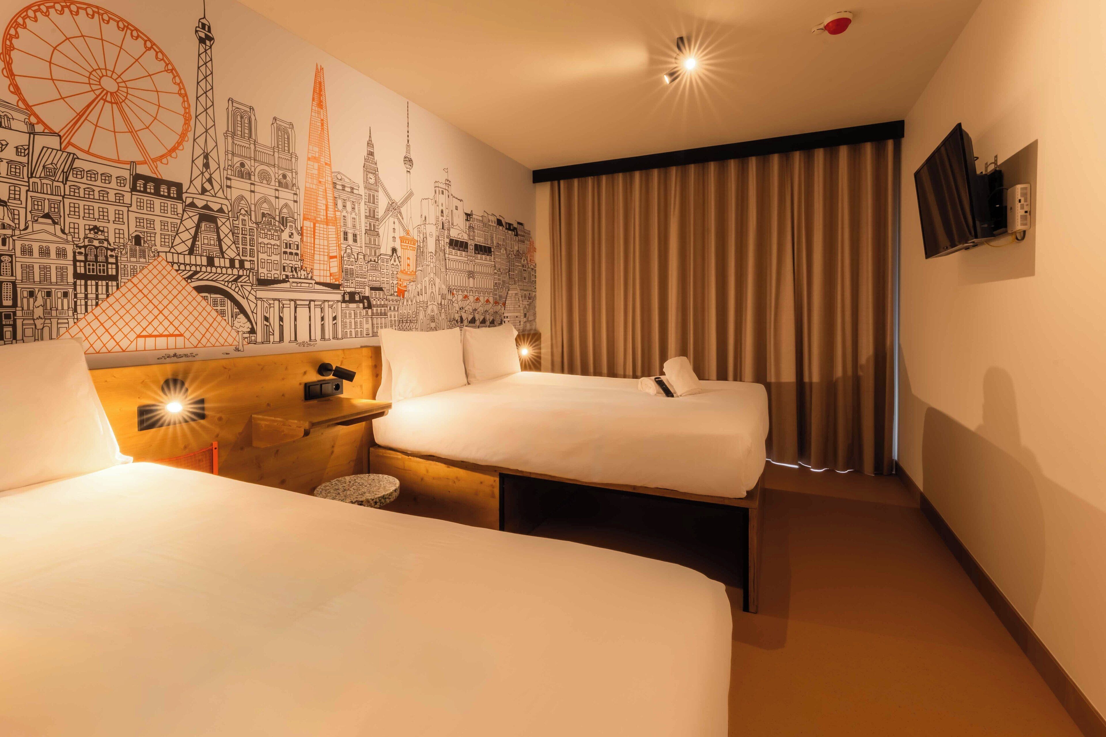 Una habitación de hotel con dos camas, un mural de paisaje urbano, un televisor y una ventana con cortinas.