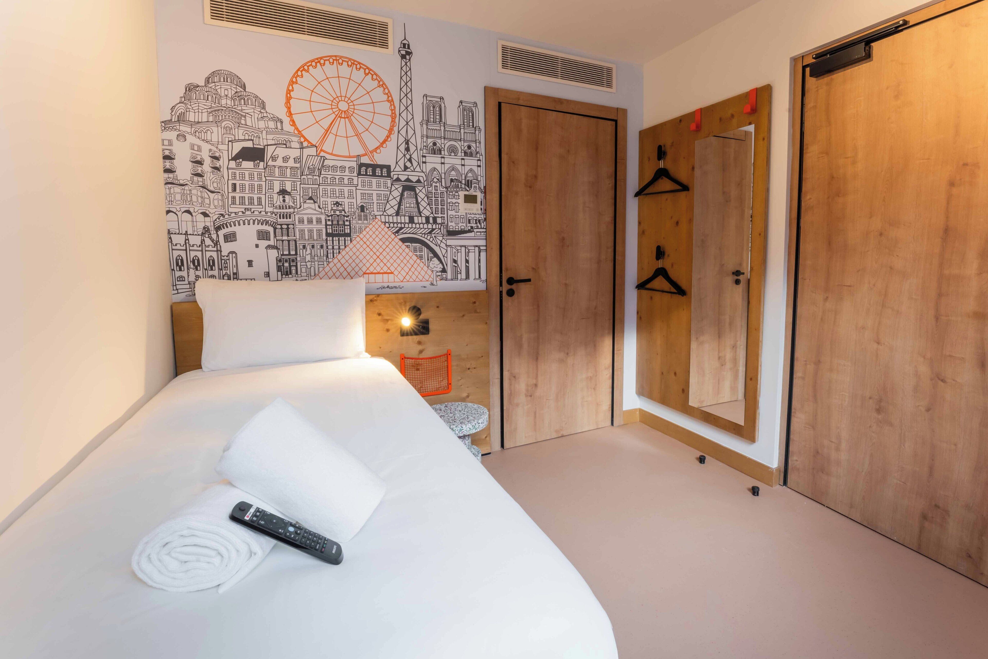 A hotel room with a bed, a remote control, a rolled towel, a wall mural, and wooden doors.