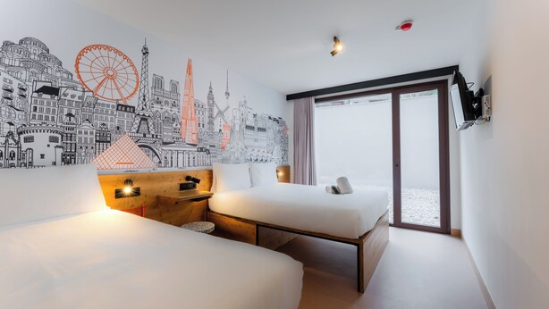 Ein Hotelzimmer mit zwei Betten, einem Stadtbild-Wandgemälde, einem großen Fenster und einer Schiebetür.