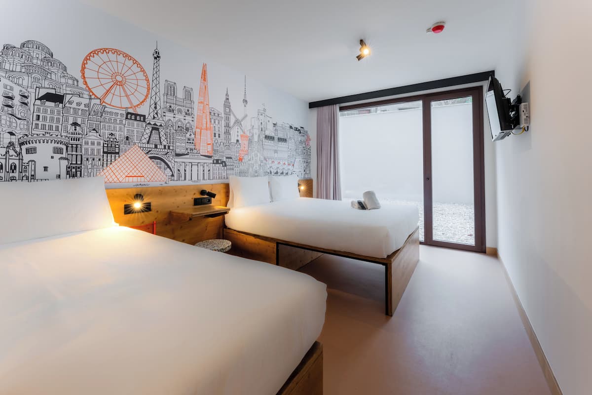 A hotel room with two beds, a cityscape mural, a large window, and a sliding door.