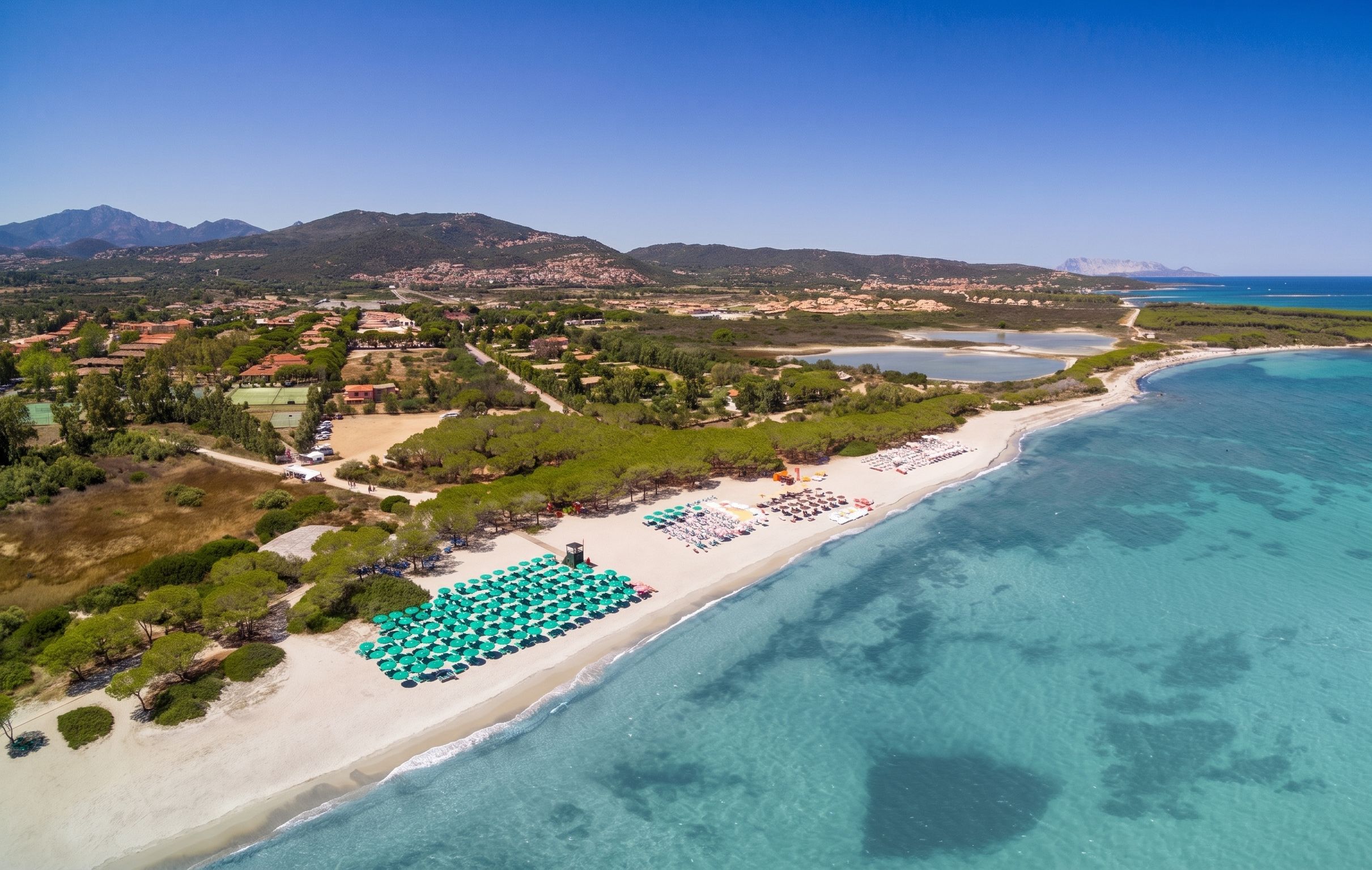 Vista aerea di una zona costiera con spiaggia, acque blu cristalline e montagne sullo sfondo.