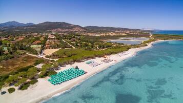Vista aerea di una zona costiera con spiaggia, acque blu cristalline e montagne sullo sfondo.