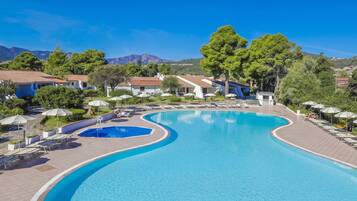 Una piscina dalla forma sinuosa, circondata da lettini e ombrelloni, con edifici e alberi sullo sfondo.