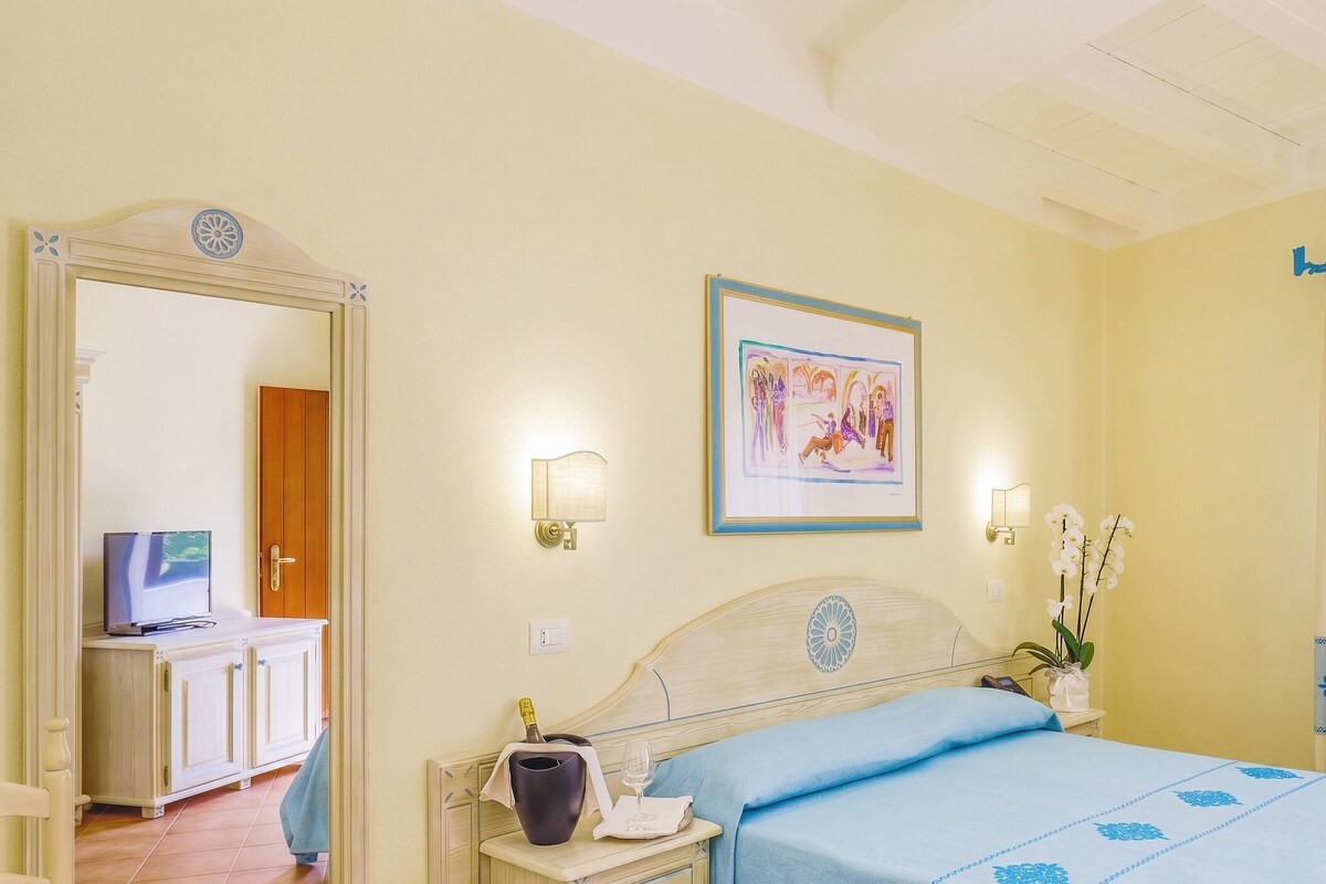 Una habitación de hotel con una cama, una mesita de noche con una botella y un vaso, un espejo, un cuadro enmarcado en la pared y una ventana con cortinas.
