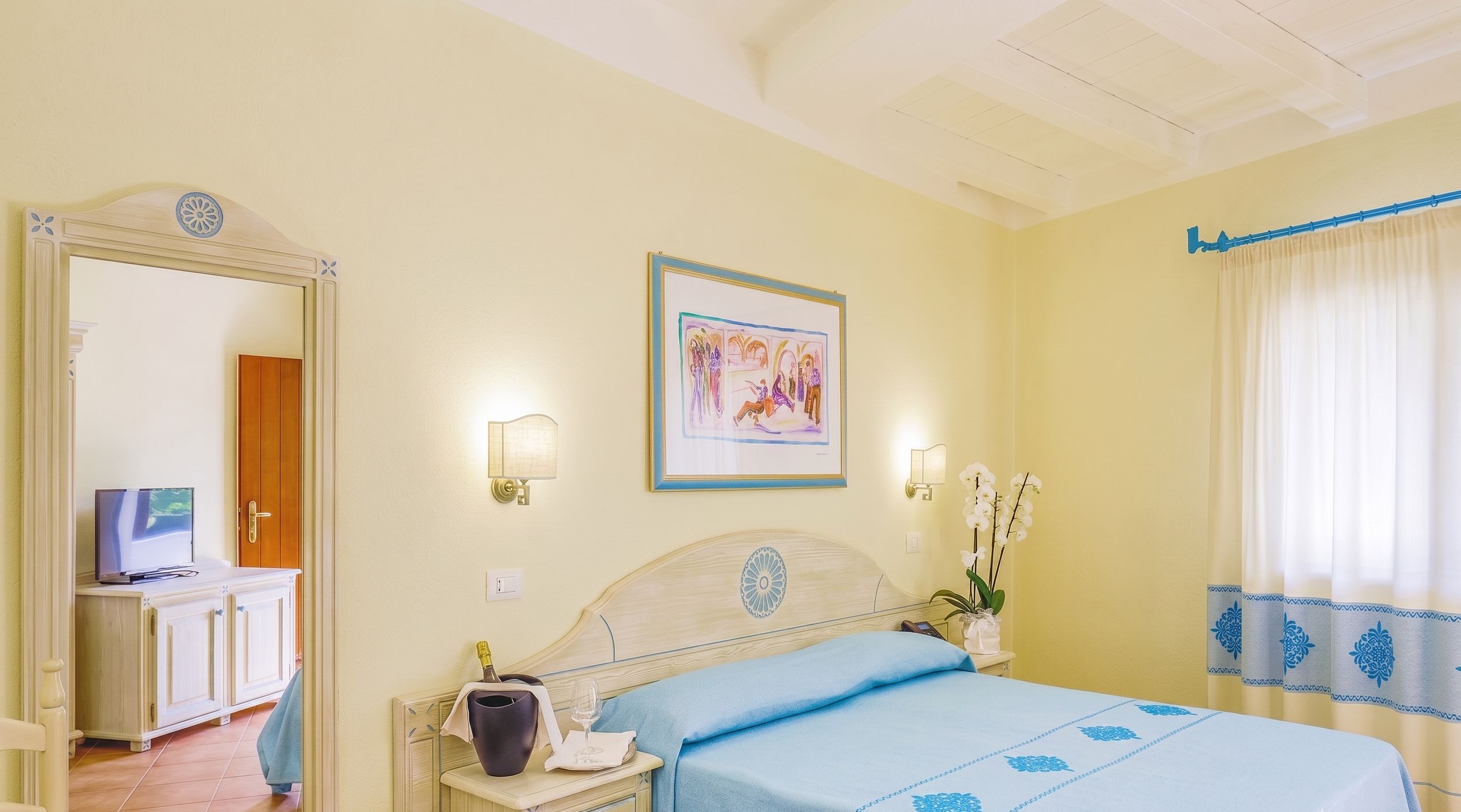 Camera d'albergo con un letto, un comodino con una bottiglia e un bicchiere, uno specchio, un quadro appeso al muro e una finestra con tende.