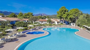 A swimming pool with a curved design, surrounded by lounge chairs and umbrellas, with buildings and trees in the background. - Li Cucutti (Sassari)