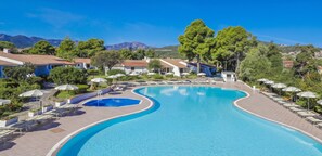 A swimming pool with a curved design, surrounded by lounge chairs and umbrellas, with buildings and trees in the background. - Li Cucutti (Sassari)