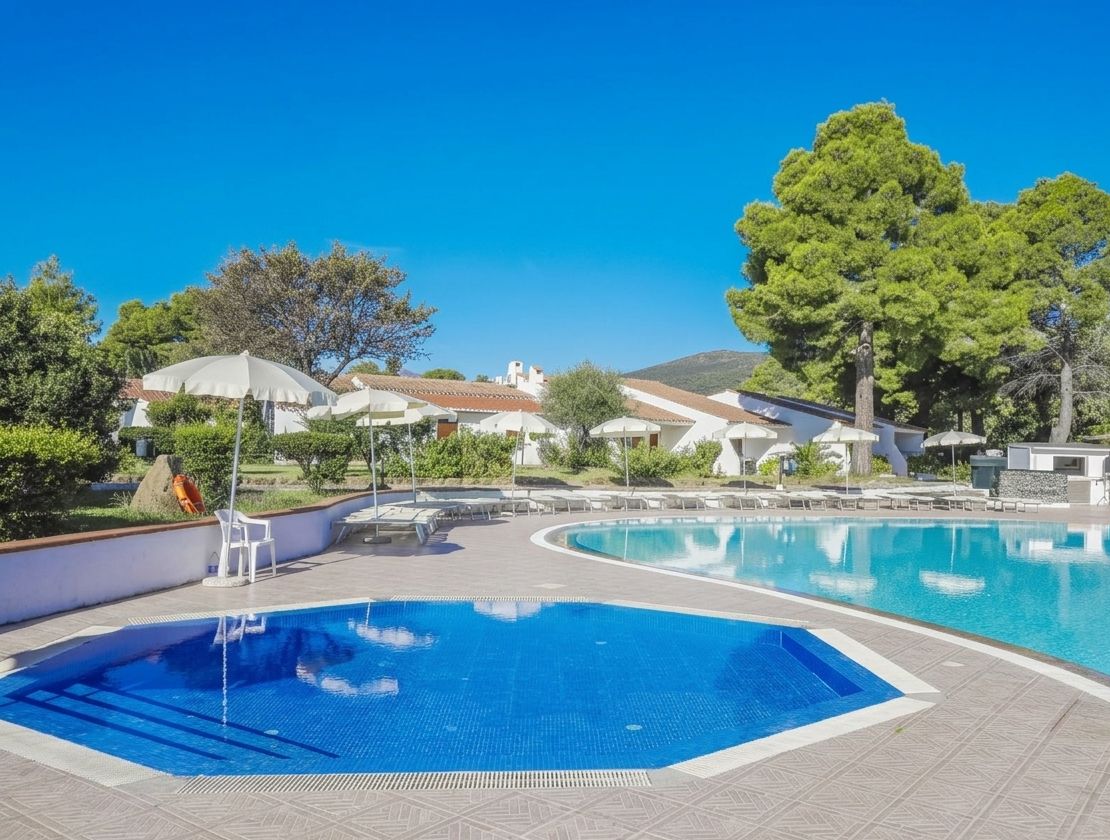 Una piscina con acqua cristallina, circondata da lettini e ombrelloni, con edifici e alberi sullo sfondo.