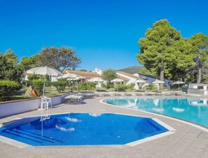 Una piscina con agua azul cristalina, rodeada de sillas de descanso y sombrillas, con edificios y árboles al fondo.