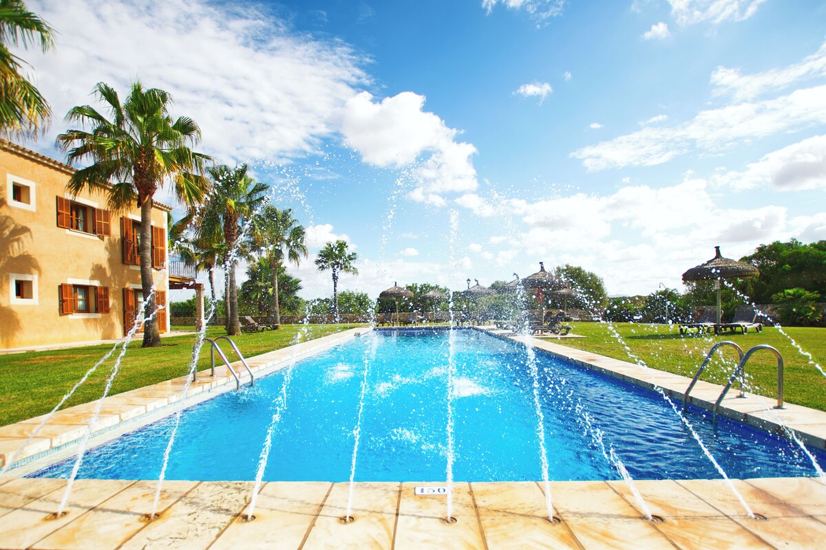 A swimming pool with water jets, surrounded by palm trees and a building with balconies.