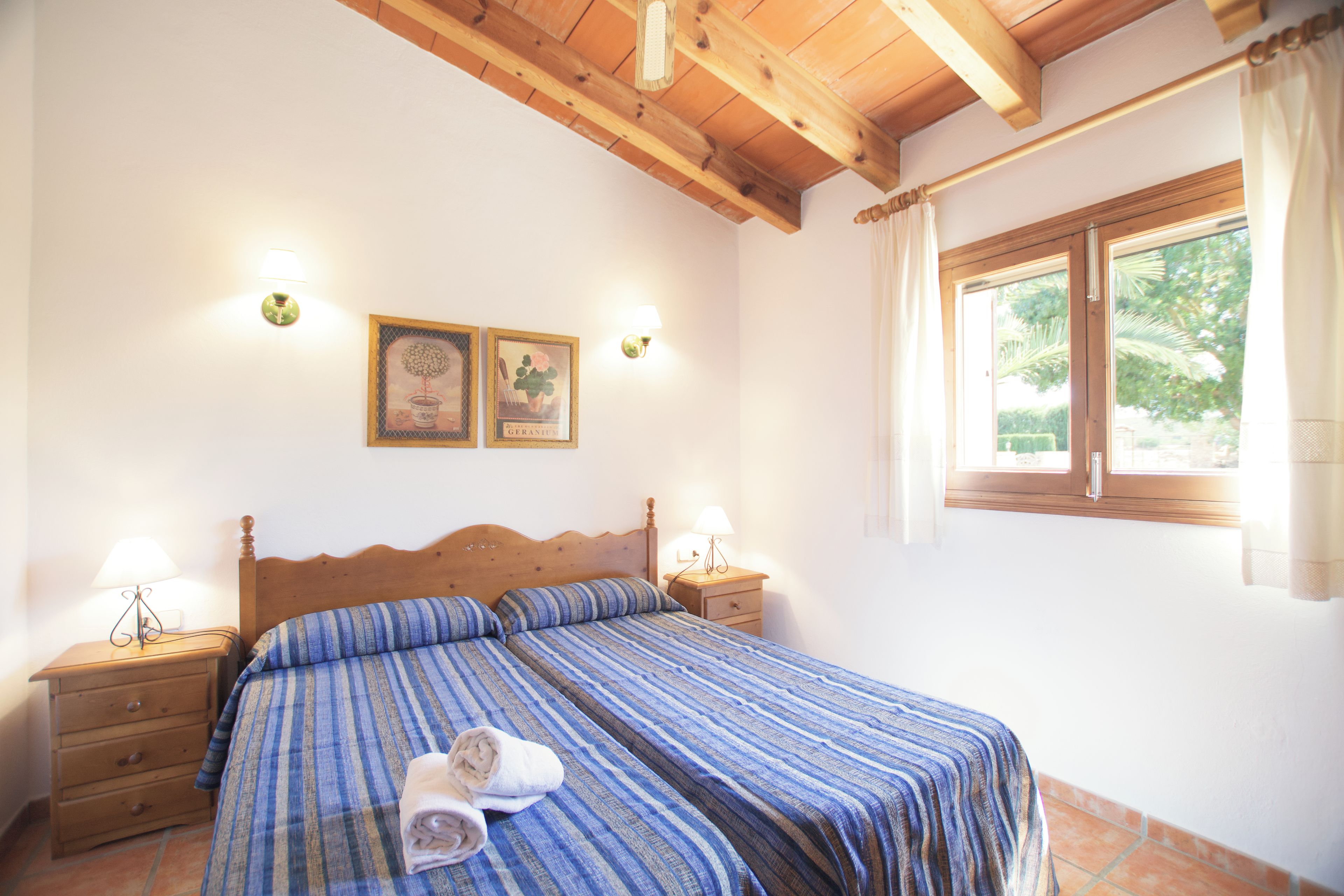 A bedroom with a wooden bed, two bedside tables with lamps, a window with curtains, and two wall-mounted light fixtures.