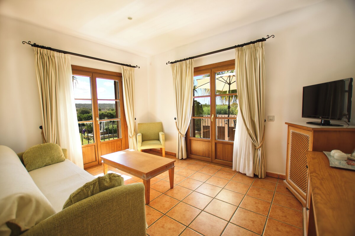 A room with a bed, a green armchair, a wooden table, a TV, and large windows with curtains.