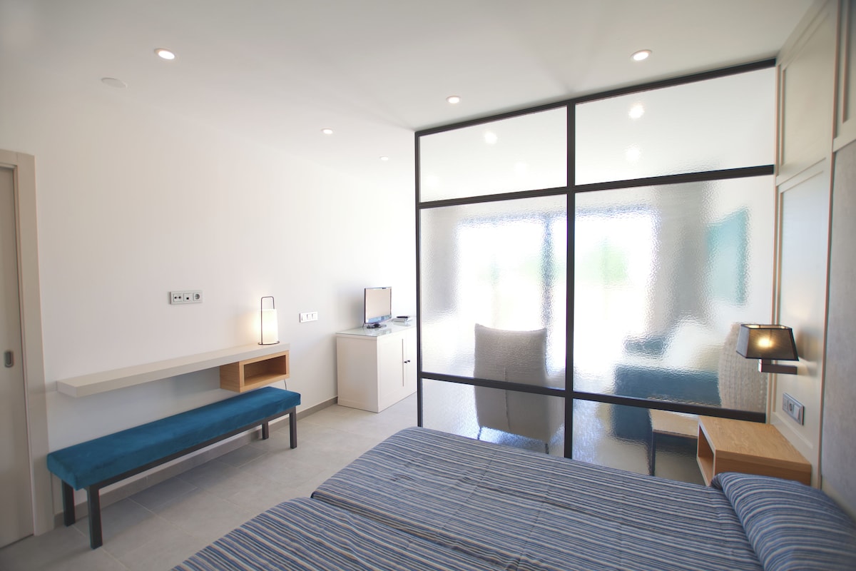 A modern hotel room with a large bed, a bench, a small desk, and a large window.