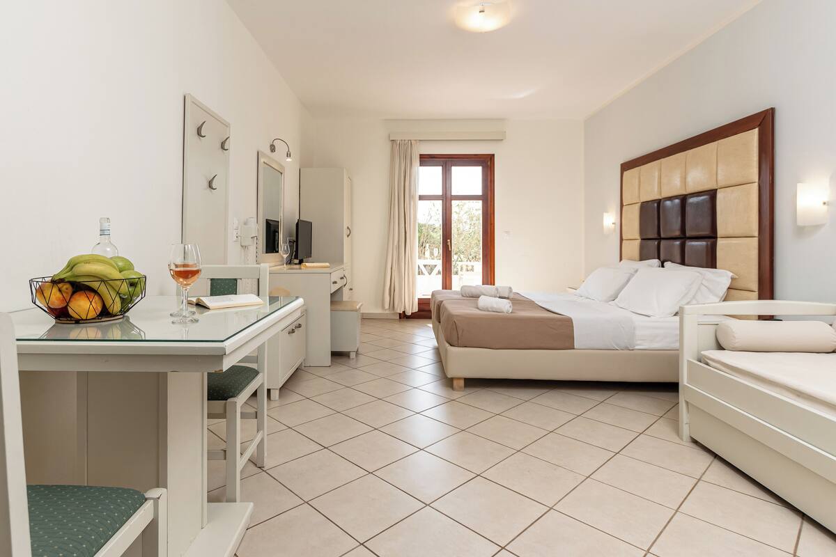 Ein Hotelzimmer mit einem Bett, einem Schreibtisch mit Stuhl, einem kleinen Tisch mit Obst und einem Glas sowie einem Fernseher.