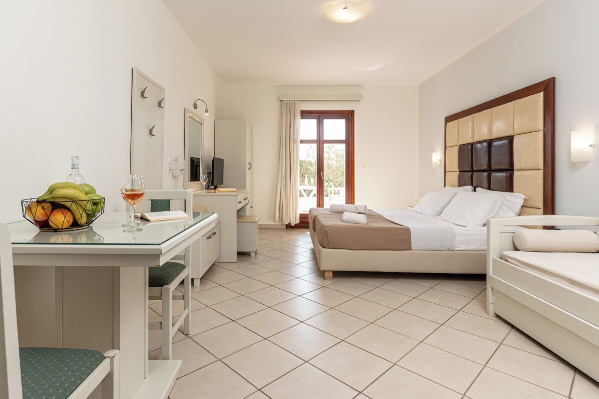 A hotel room with a bed, a desk with a chair, a small table with fruit and a glass, and a television.