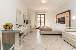 A hotel room with a bed, a desk with a chair, a small table with fruit and a glass, and a television.