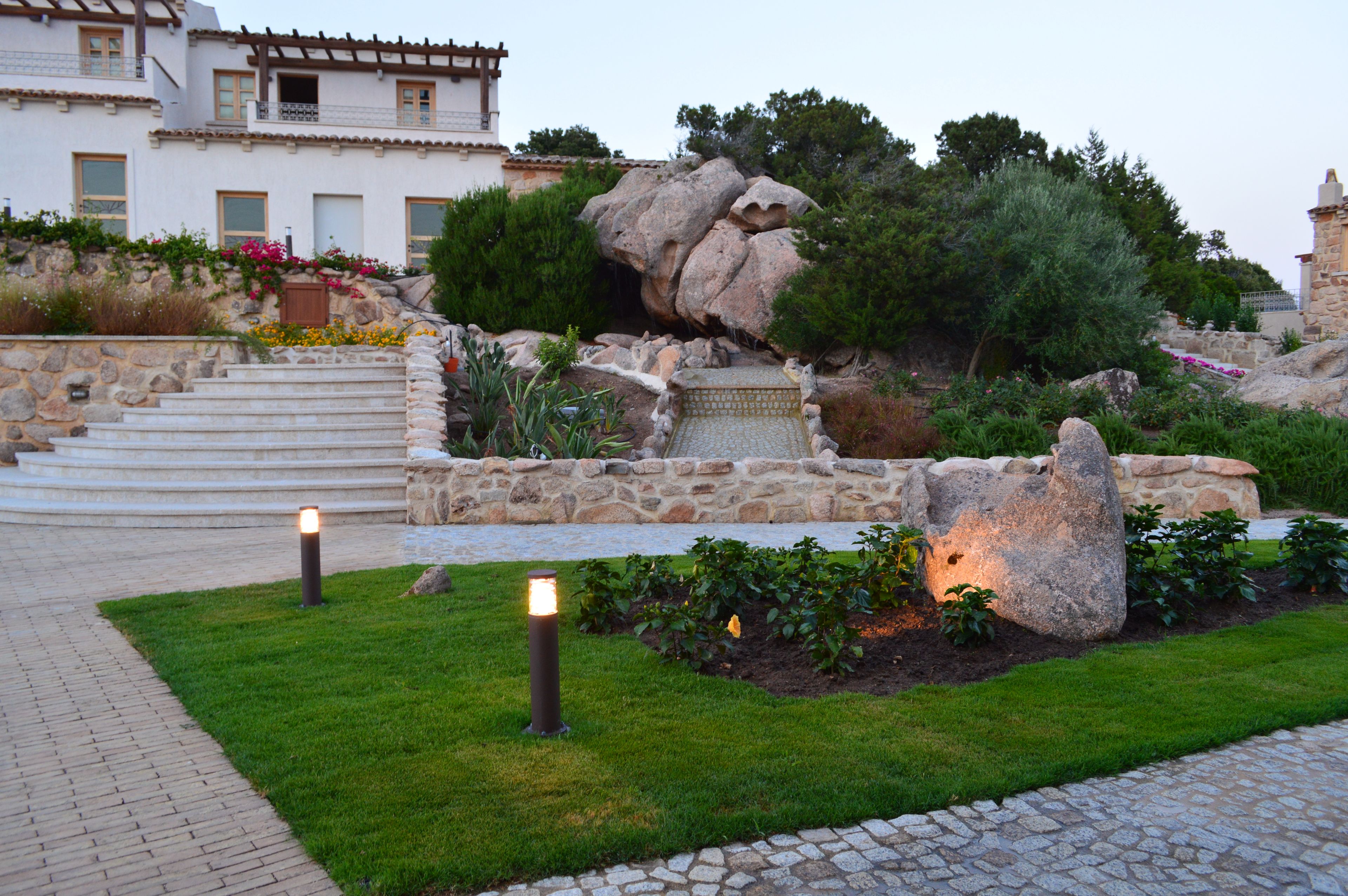 A landscaped garden with a stone pathway, a rock formation, and a building with multiple windows and balconies.