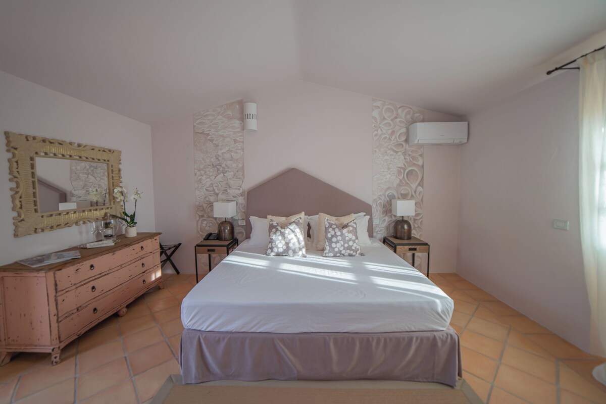 A bedroom with a large bed, two bedside tables, a dresser, a mirror, and an air conditioning unit.