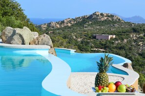 A swimming pool with a curved edge, surrounded by rocks and pebbles. In the foreground, there is a plate with various fruits including a pineapple, grapes, and apples. The background features a mountainous landscape with greenery and a building. - Hotel Borgo Smeraldo (Olbia)