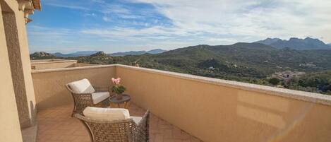 Un balcón con mobiliario de mimbre y una mesita, con vistas a un paisaje montañoso.