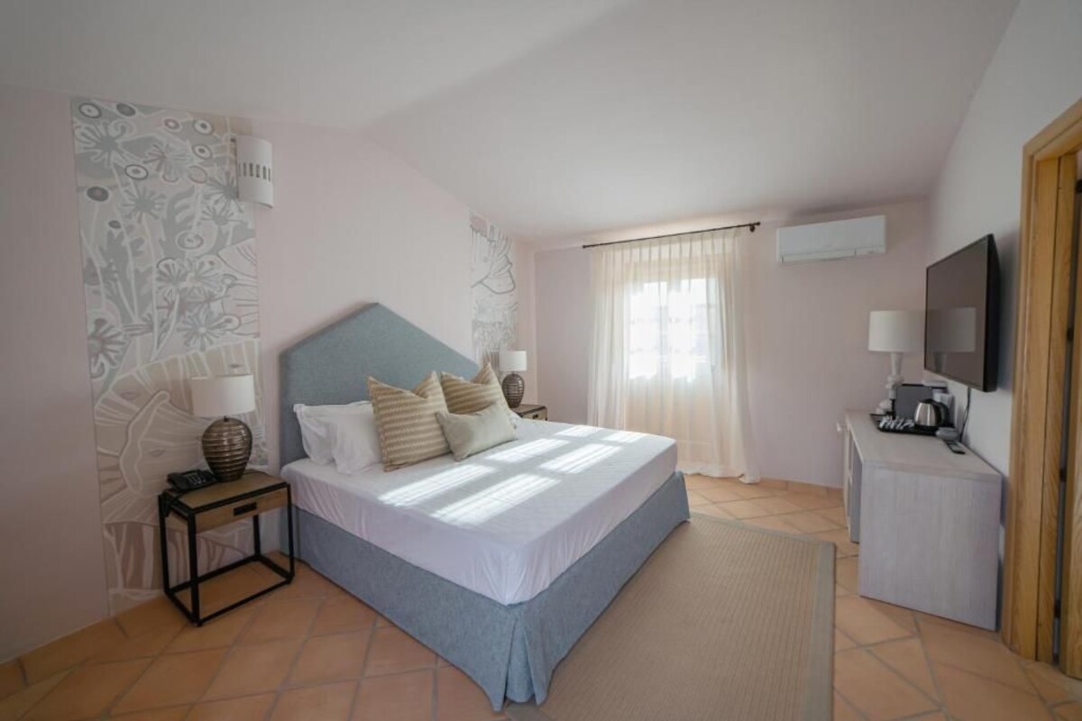 A bedroom with a bed, bedside table, lamp, TV, and a small desk.