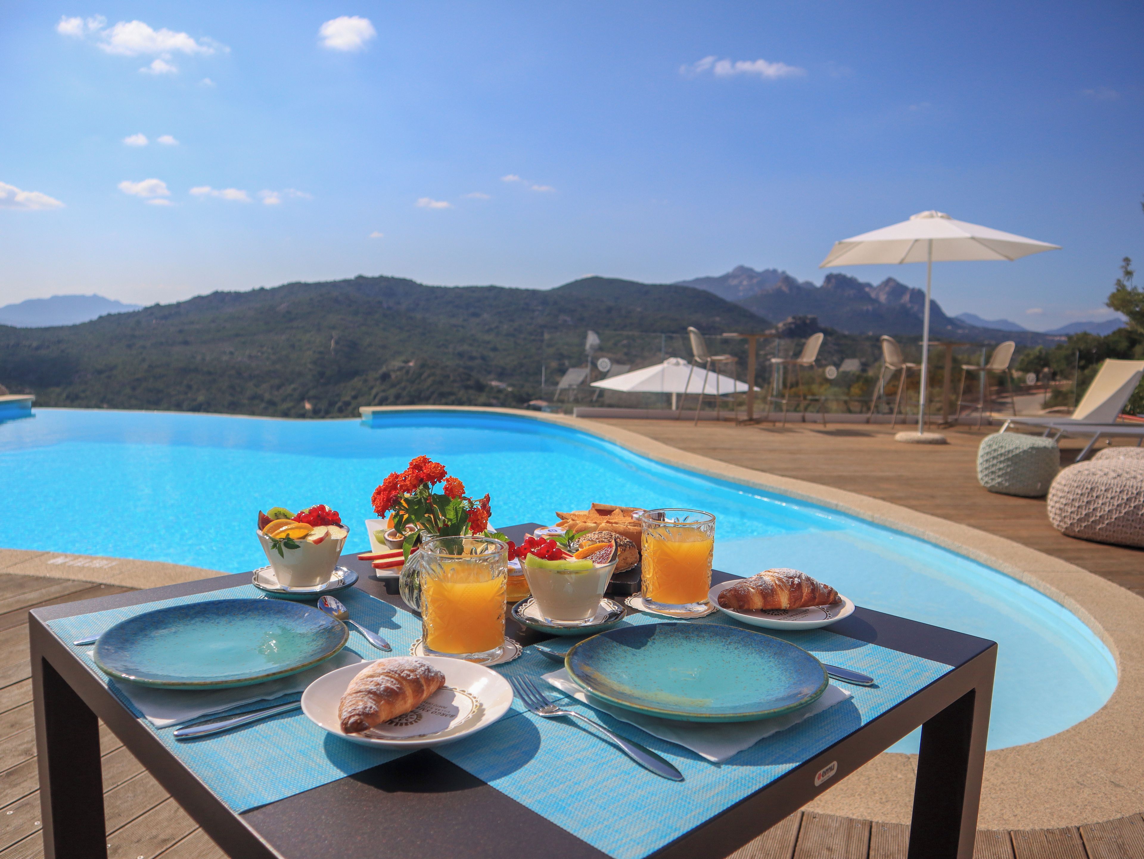 A dining table with plates, cutlery, and a glass of orange juice, set against a poolside view with mountains in the background.
