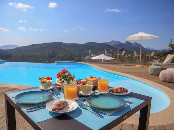 A dining table with plates, cutlery, and a glass of orange juice, set against a poolside view with mountains in the background. - Hotel Borgo Smeraldo (Olbia)
