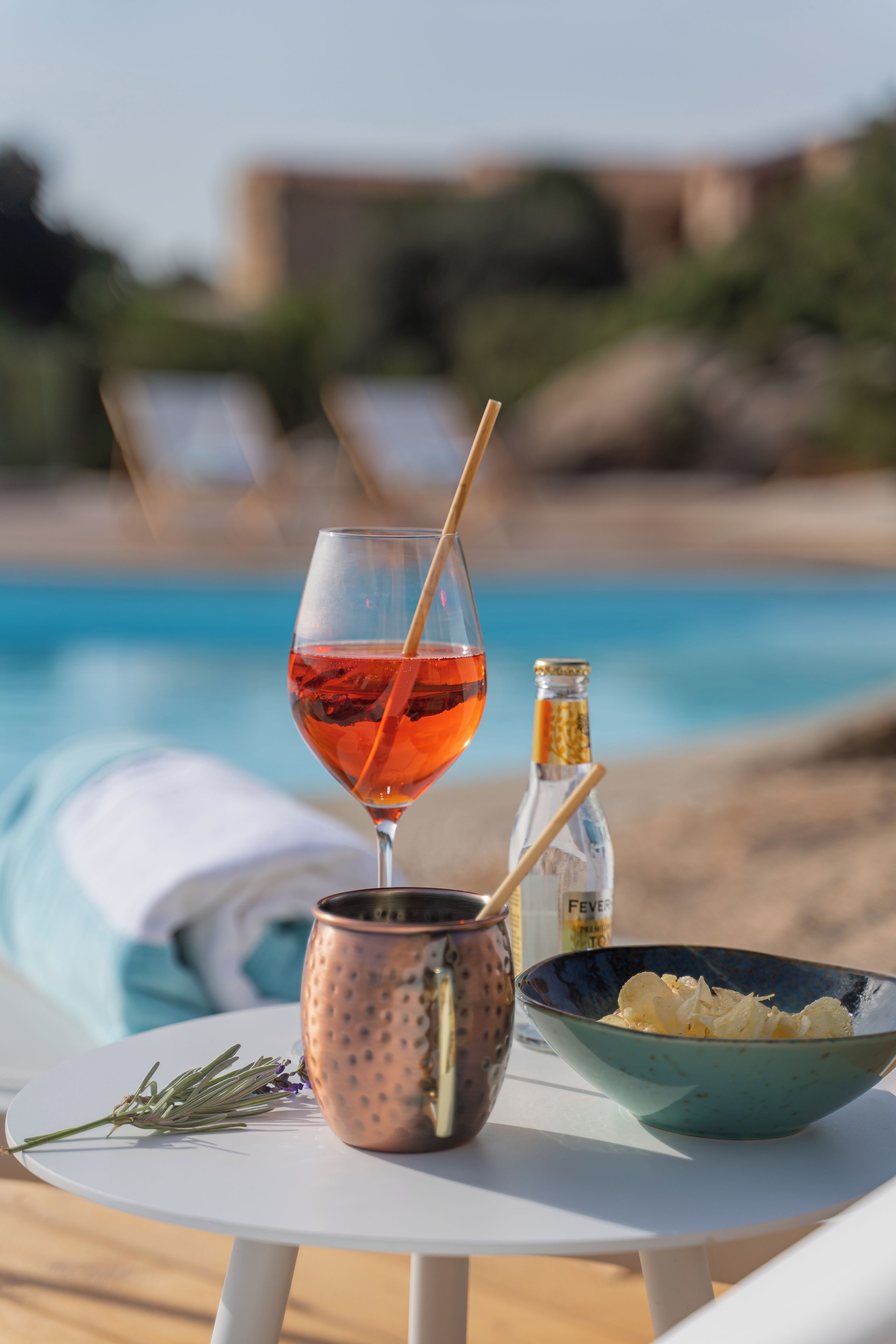 A table with a glass of red drink, a copper mug, a small bottle, and a bowl of snacks by a poolside.