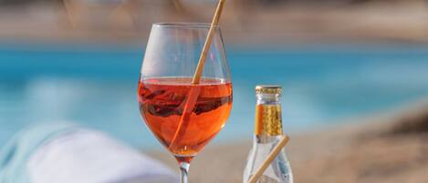 A table with a glass of red drink, a copper mug, a small bottle, and a bowl of snacks by a poolside.